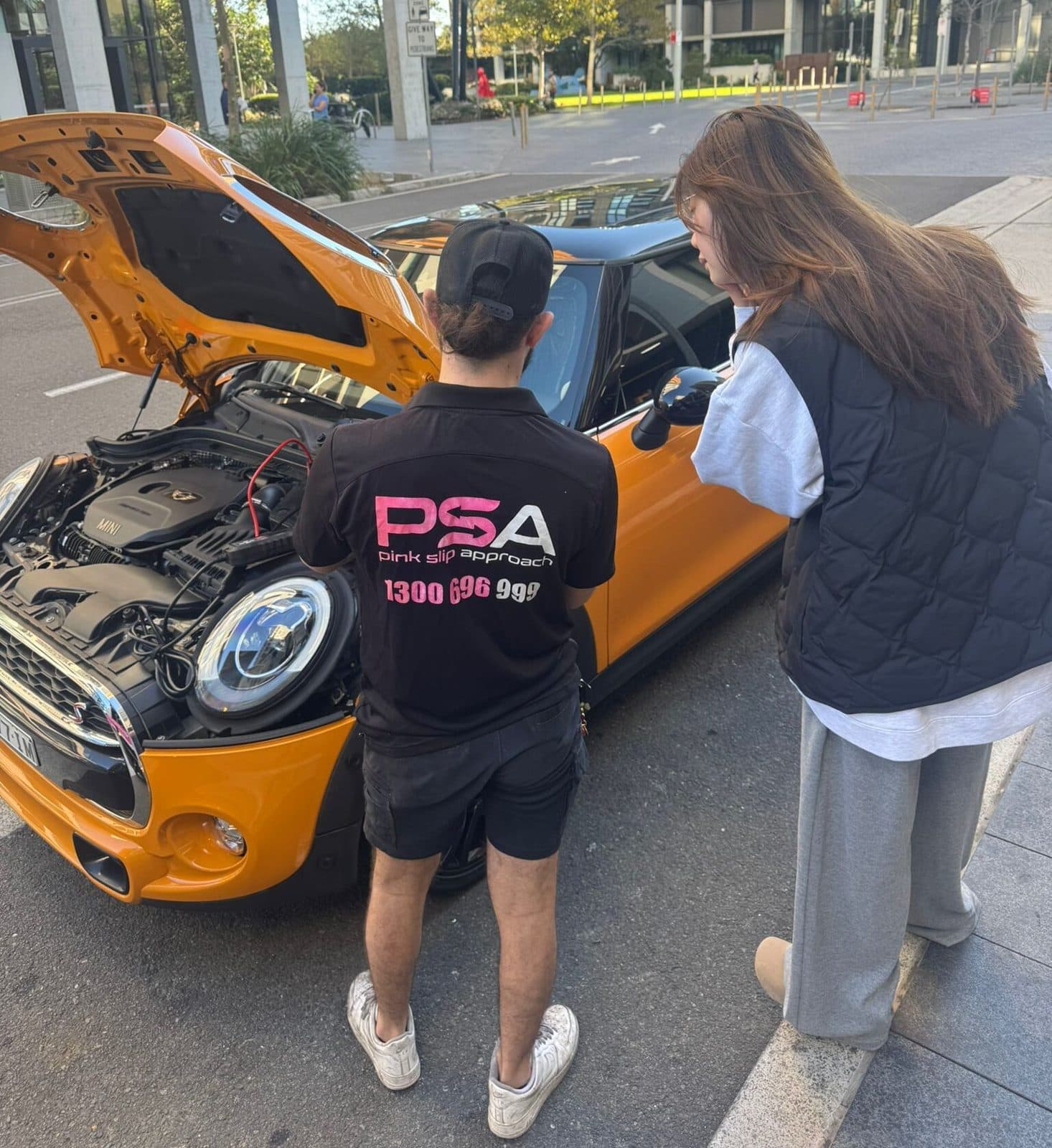 A Pink Slip Approach technician showing a customer the engine bay of an orange Mini