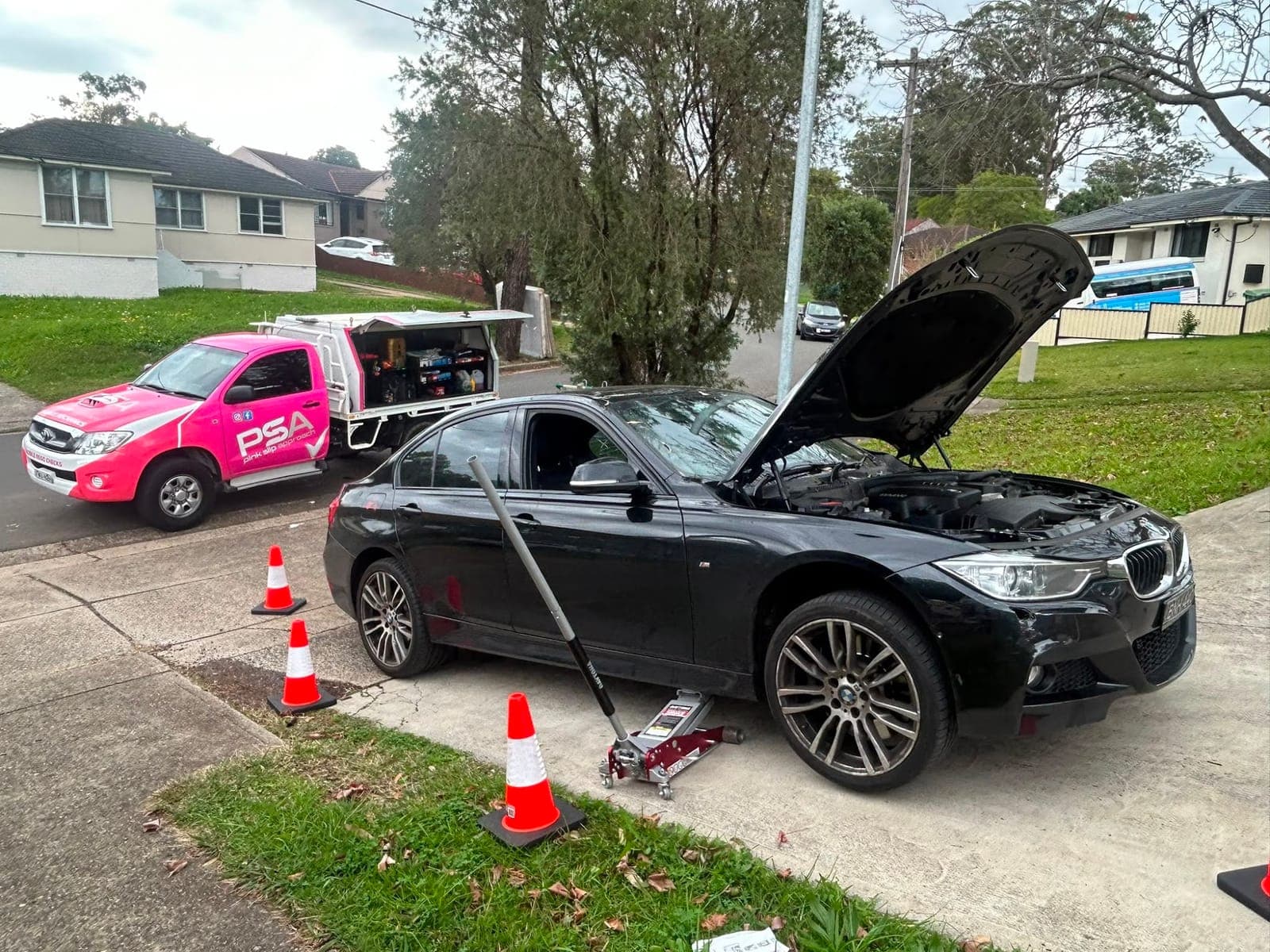 A black BMW sedan with its bonnet up and a trolley jack beside it at the kerb, a pink PSA service ute parked behind