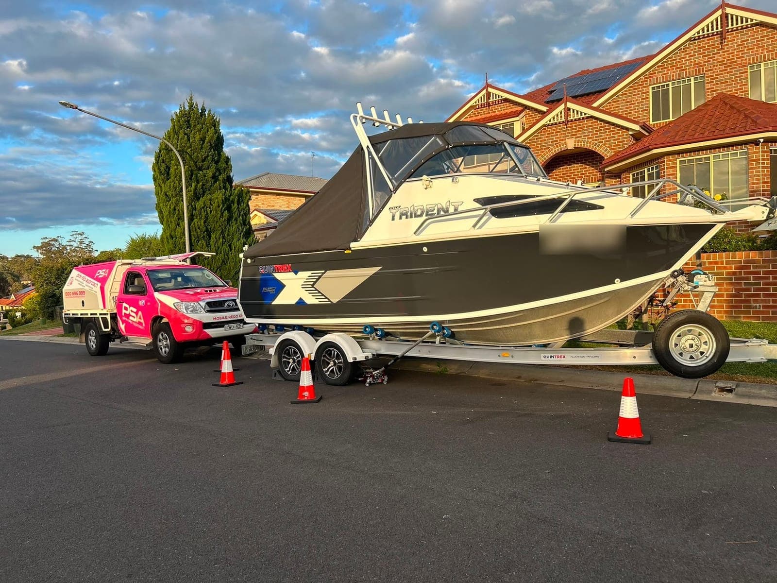 A boat on a tandem trailer in a suburban street, cones around it and a pink PSA ute behind