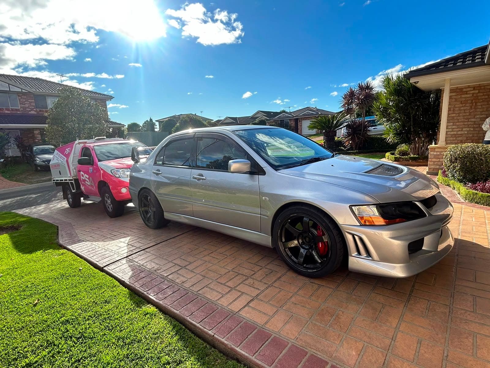 A silver Mitsubishi Lancer Evolution on a paved driveway in the sun, a pink PSA ute parked behind it