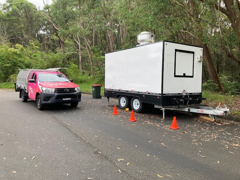 A white tandem-axle enclosed trailer beside a pink PSA service ute, with cones and wheel chocks in place