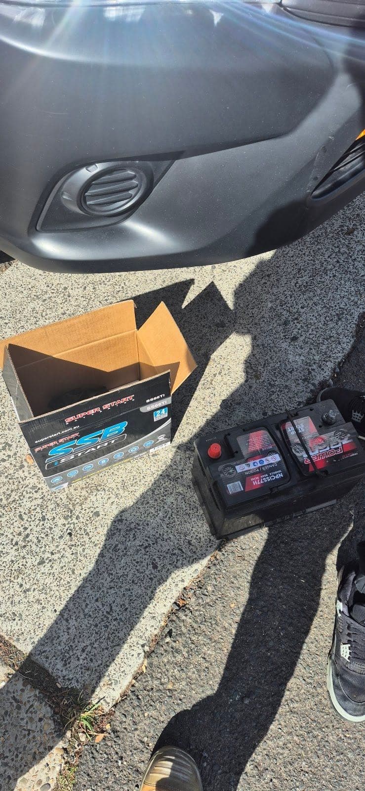 An old car battery and a new one in its box on the kerb beside a grey car