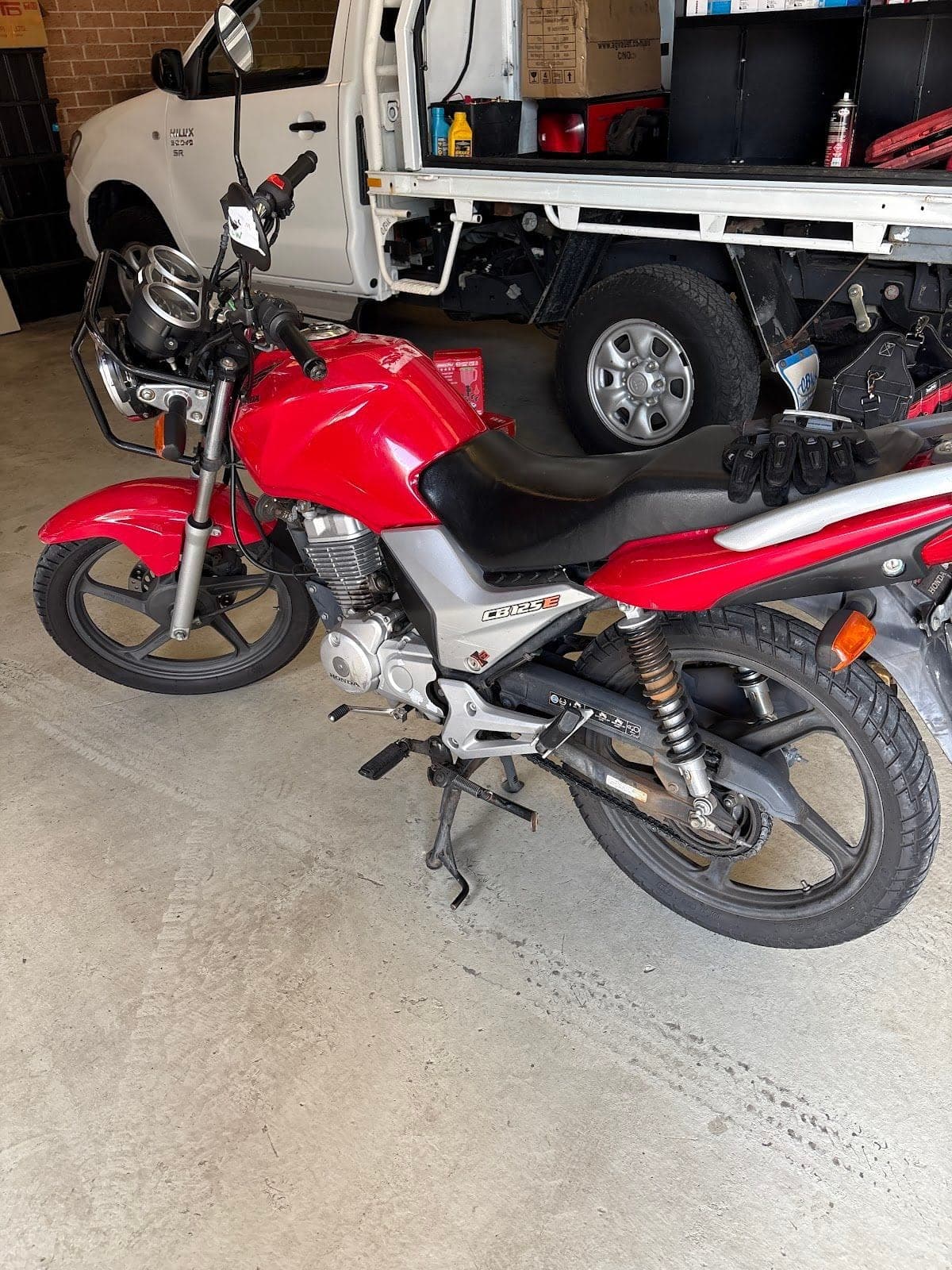 A red Honda CB125E motorbike in a garage, a white ute behind it