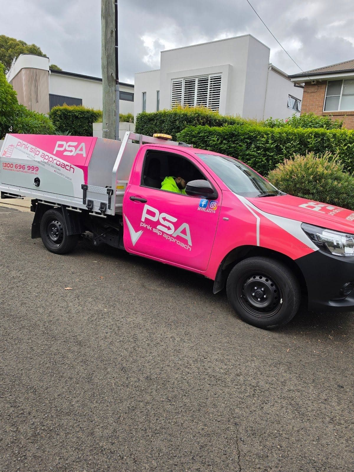 A pink PSA service ute parked outside a house with hedges, a technician in the cab