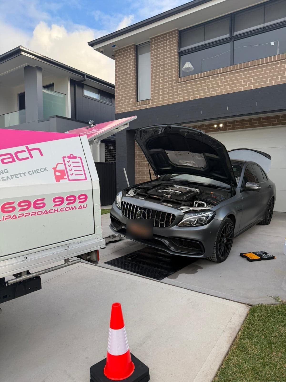 A grey Mercedes-AMG sedan with its bonnet up on a home driveway, a cone in front and a PSA ute’s open canopy beside it