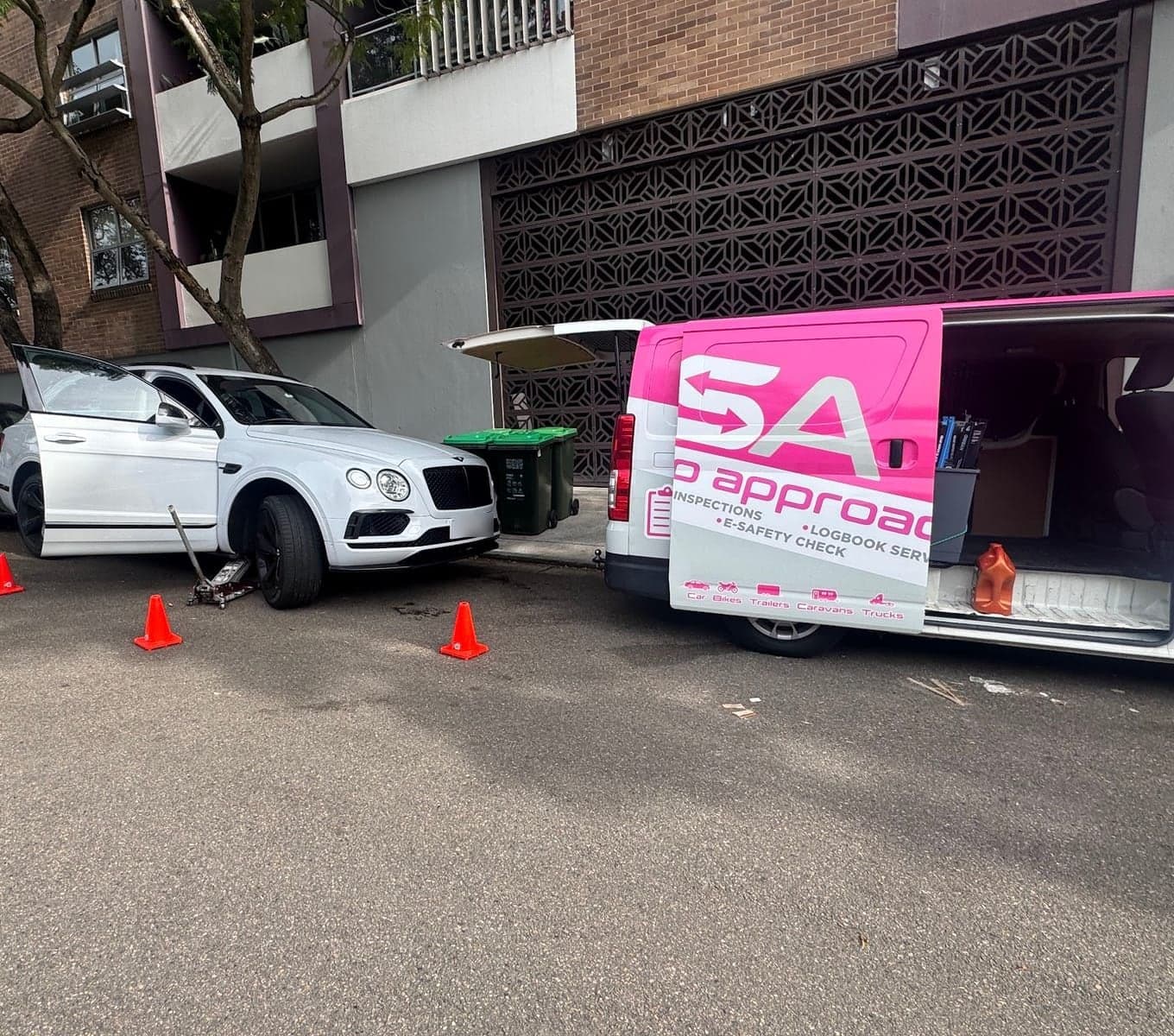 A white Bentley SUV on a jack outside an apartment block, cones around it and a PSA van with its back doors open beside it