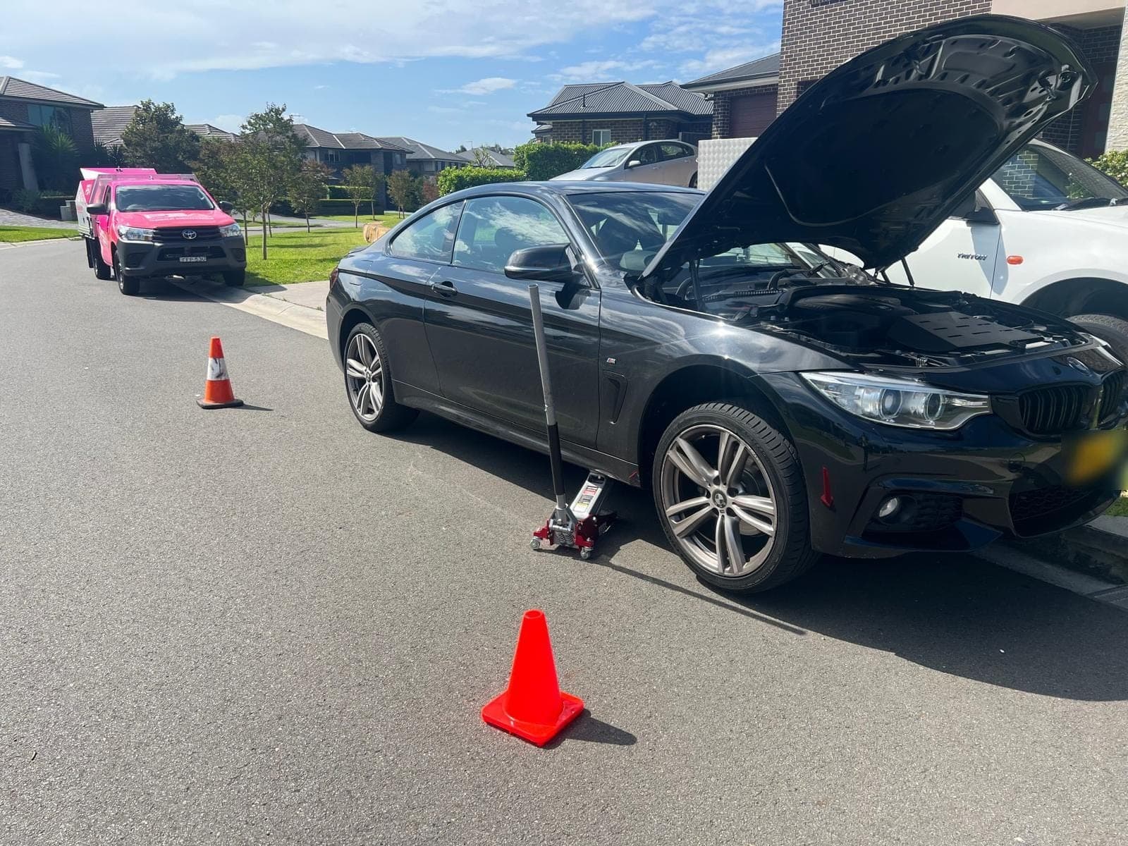 A black BMW coupe on a jack with its bonnet up in a suburban street, cones out and a pink PSA ute parked behind