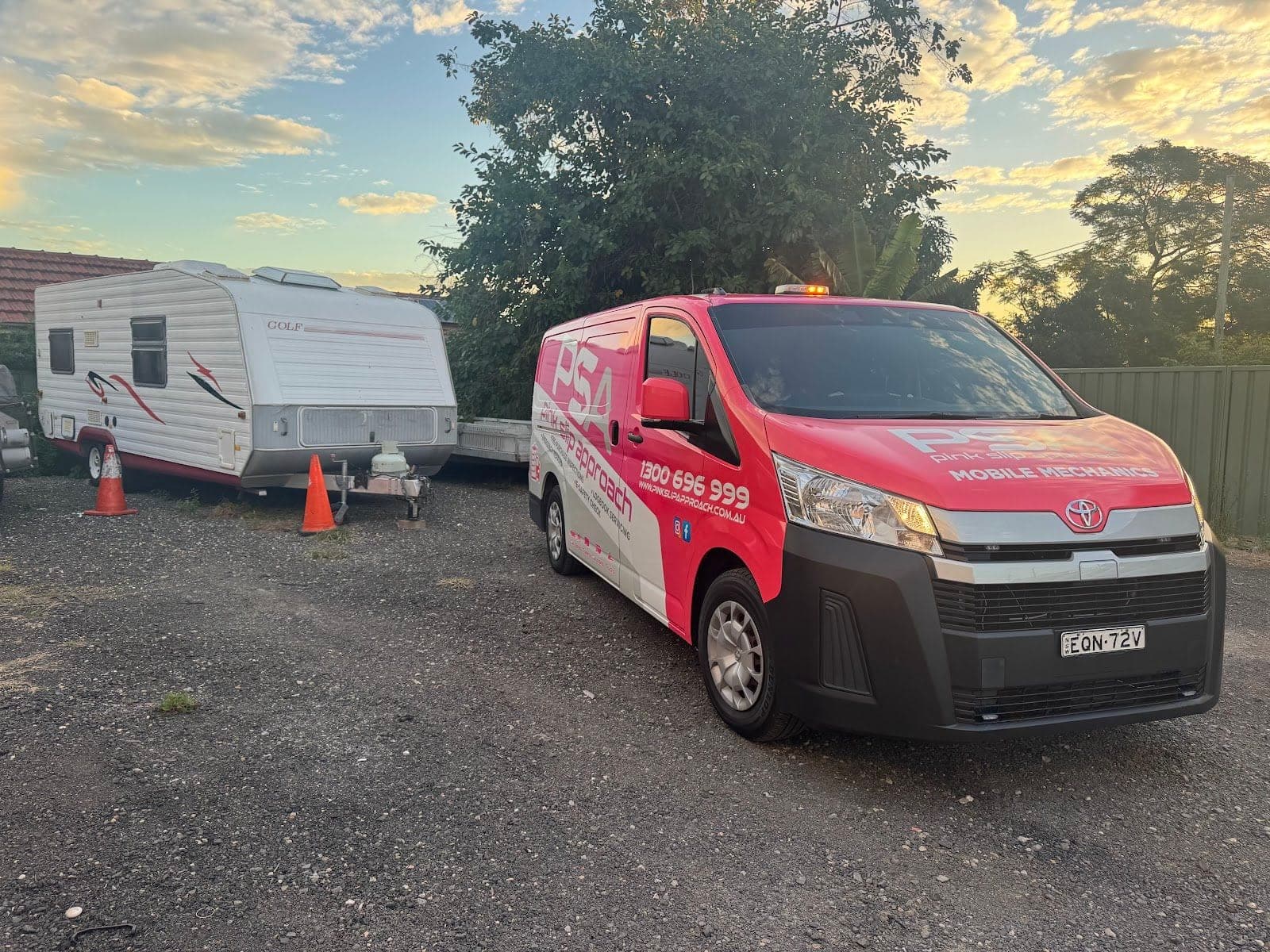 A caravan with cones beside its drawbar in a gravel yard, a pink PSA van parked next to it at sunset