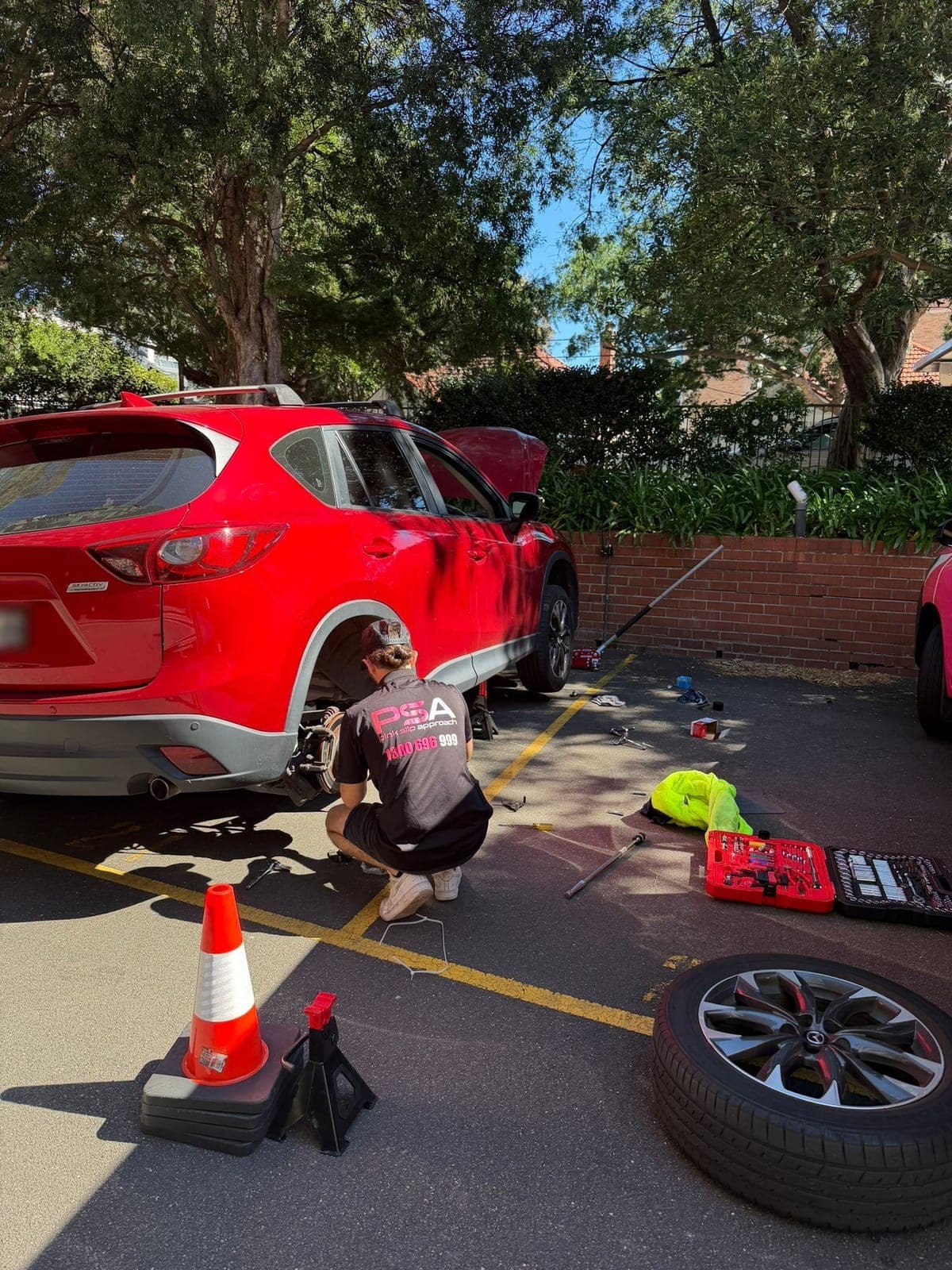 A PSA technician crouched at the rear brake of a red Mazda SUV with its wheel off, the wheel and tools laid out in a car park