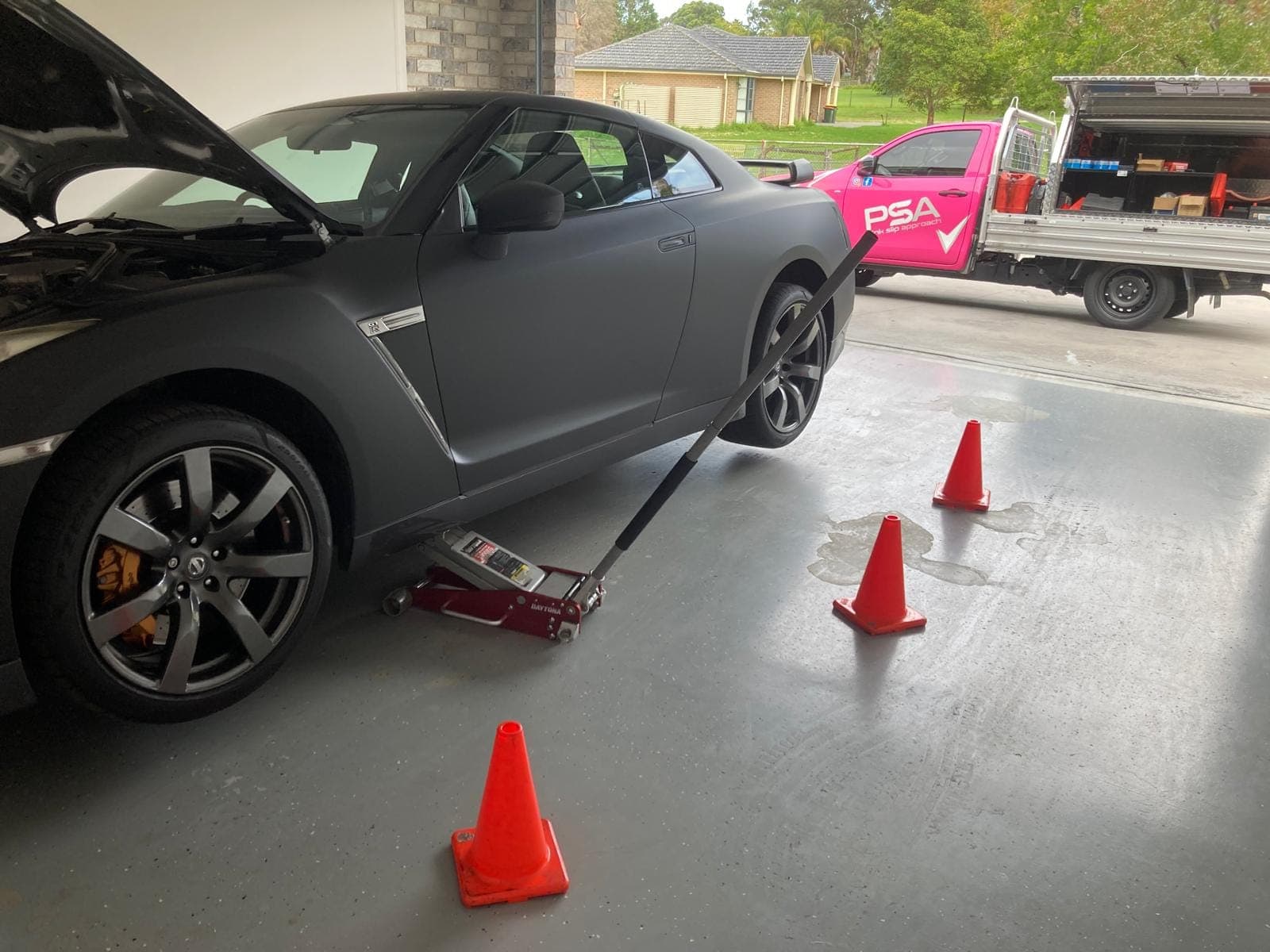 A grey Nissan GT-R on a trolley jack in a home garage, cones on the floor and a PSA ute on the driveway outside