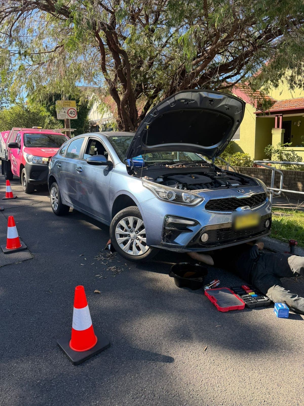 A light blue Kia hatchback with its bonnet up at the kerb, a technician underneath it, cones around it and a pink PSA ute behind