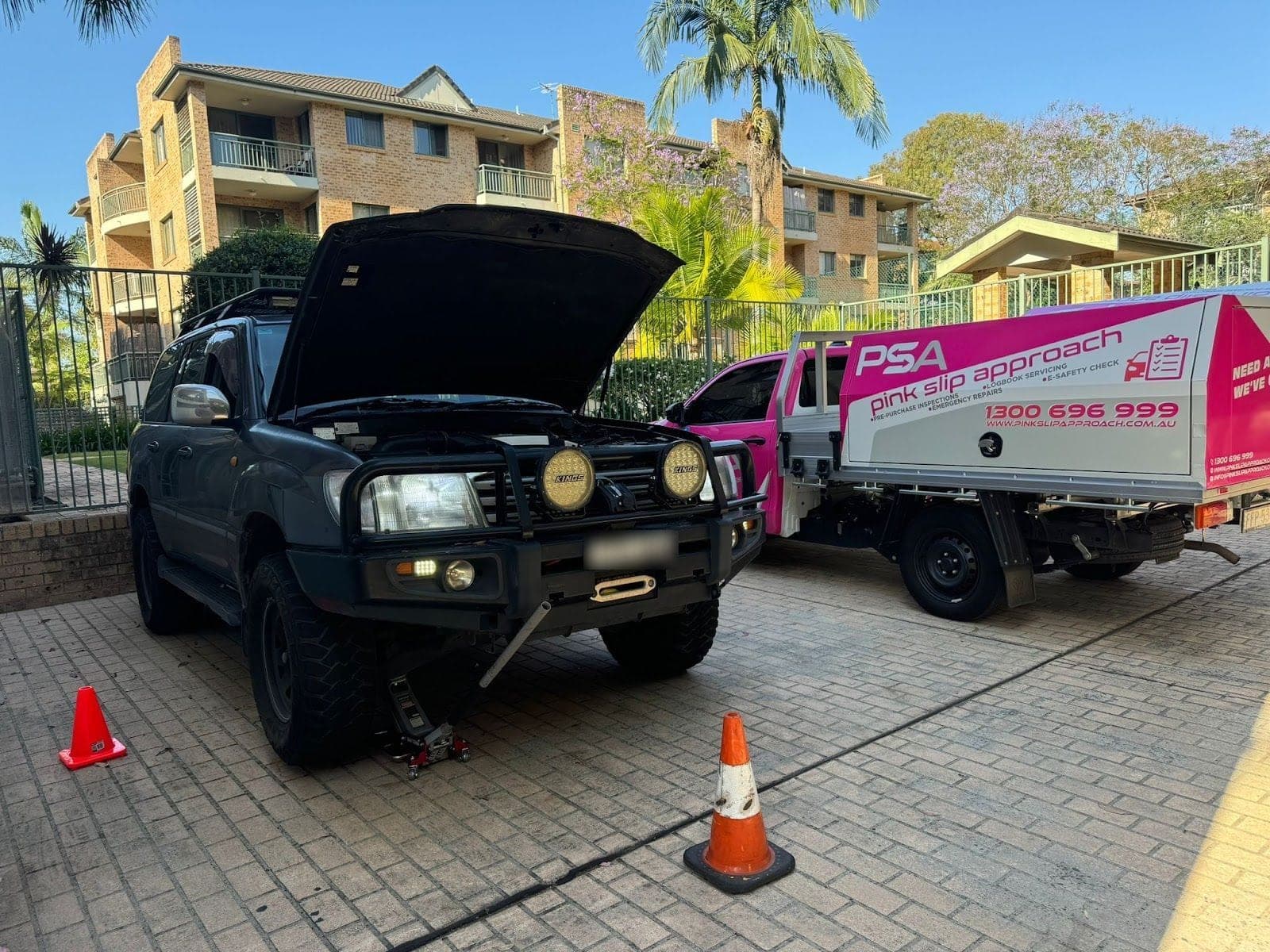 An older Toyota LandCruiser with its bonnet up on a jack in an apartment block's driveway, beside a pink PSA service ute