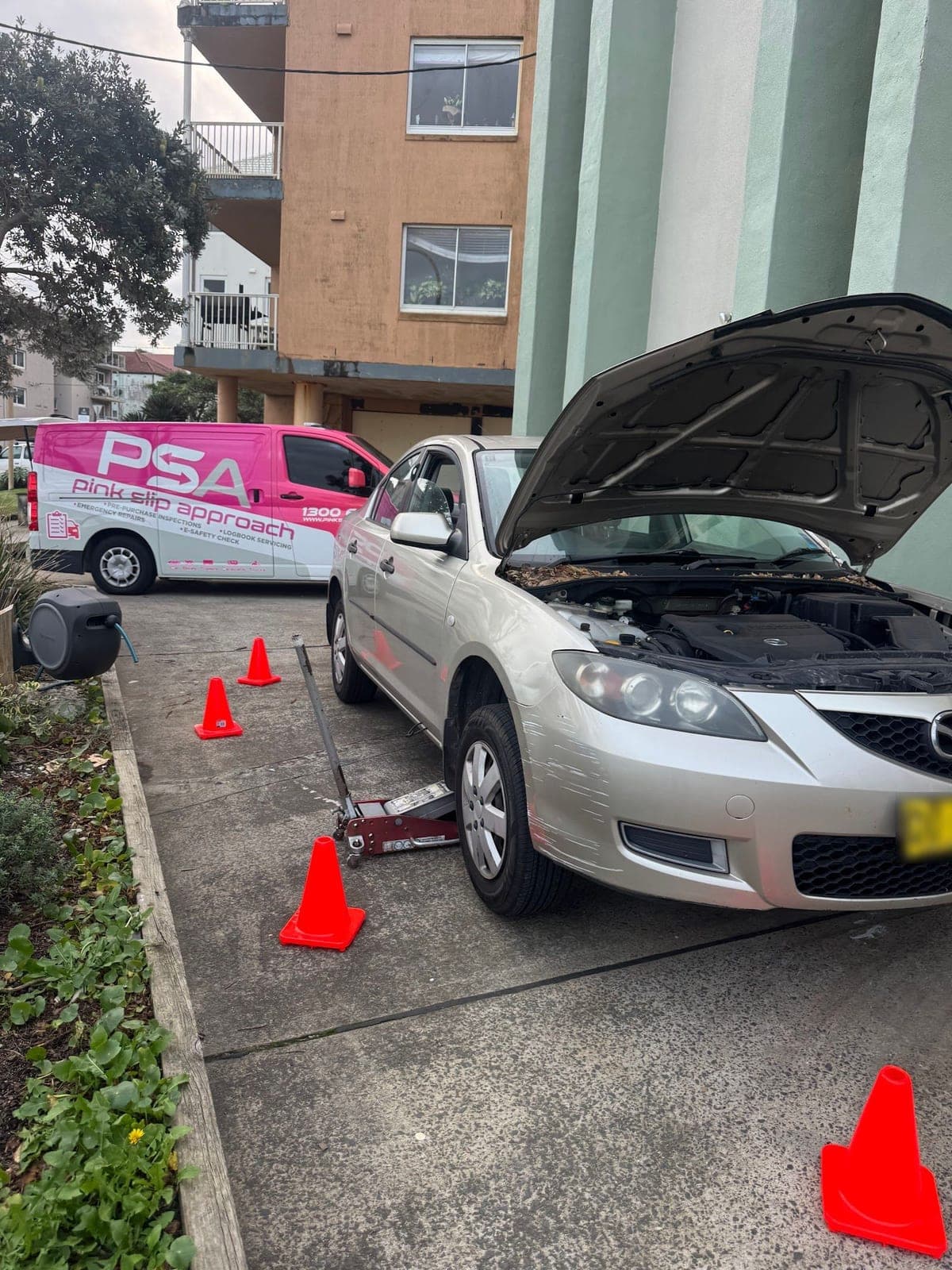 A silver Mazda3 sedan on a jack with its bonnet up in an apartment block's driveway, cones out and a PSA van behind it