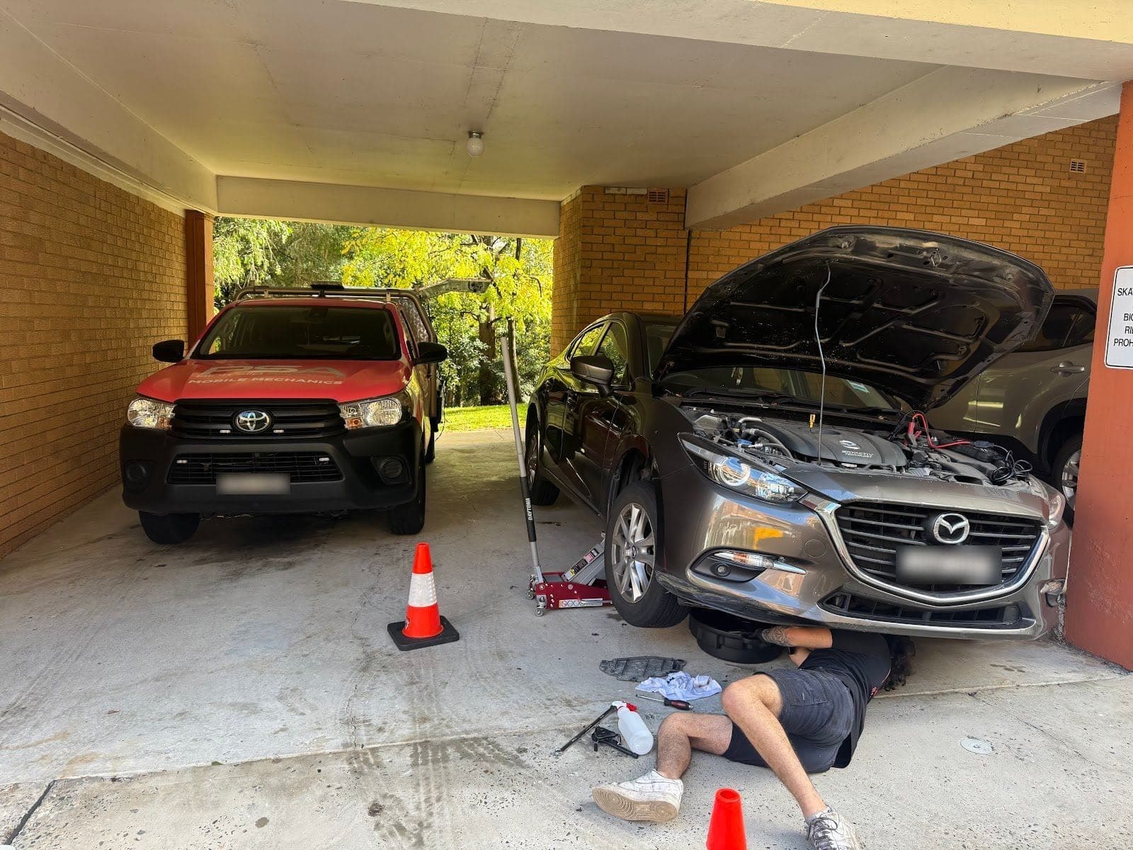 A PSA technician lying under a grey Mazda3 with its bonnet up in a carport, a red PSA ute parked beside it