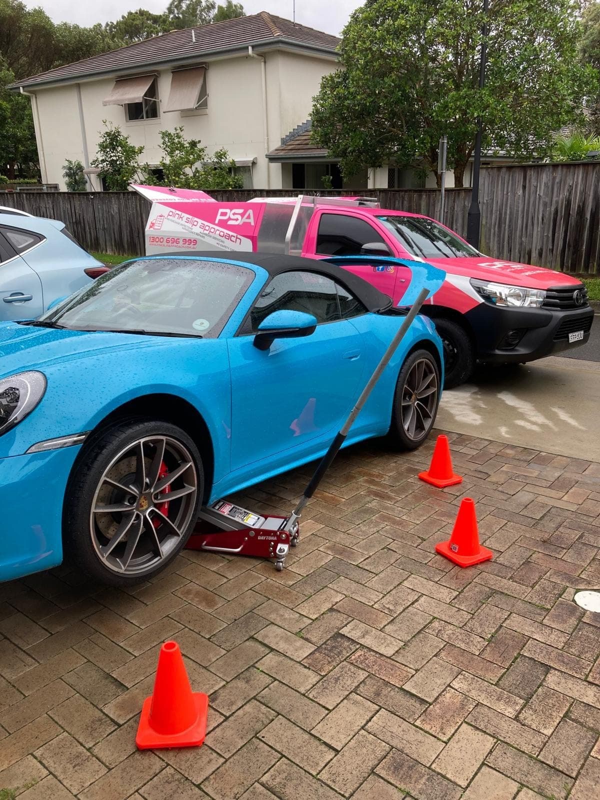 A blue Porsche 911 on a trolley jack on a paved driveway, cones around it and a pink PSA ute behind