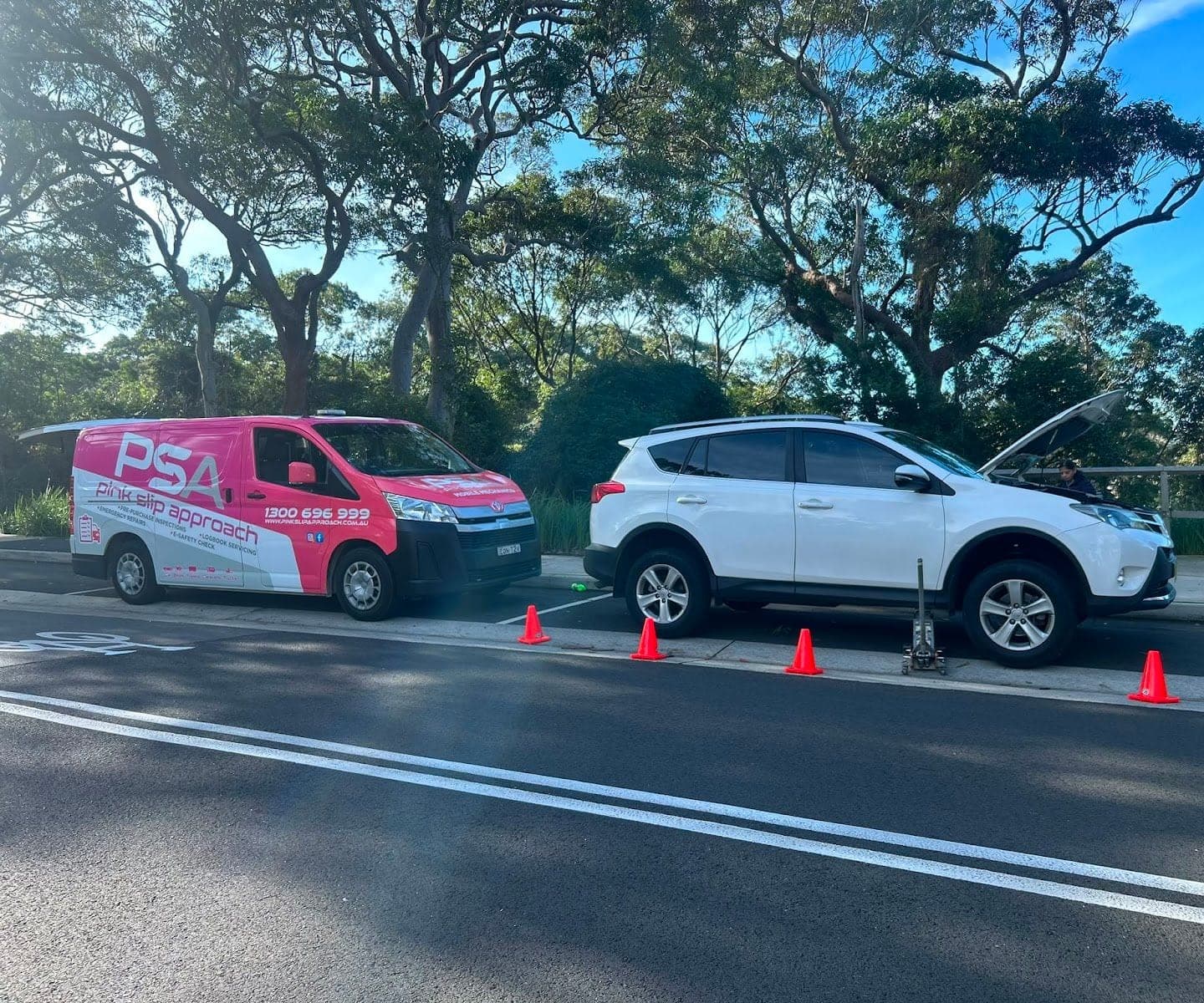 A white Toyota RAV4 on a jack at the kerb with its bonnet up, cones around it and a pink PSA van parked in front, under gum trees