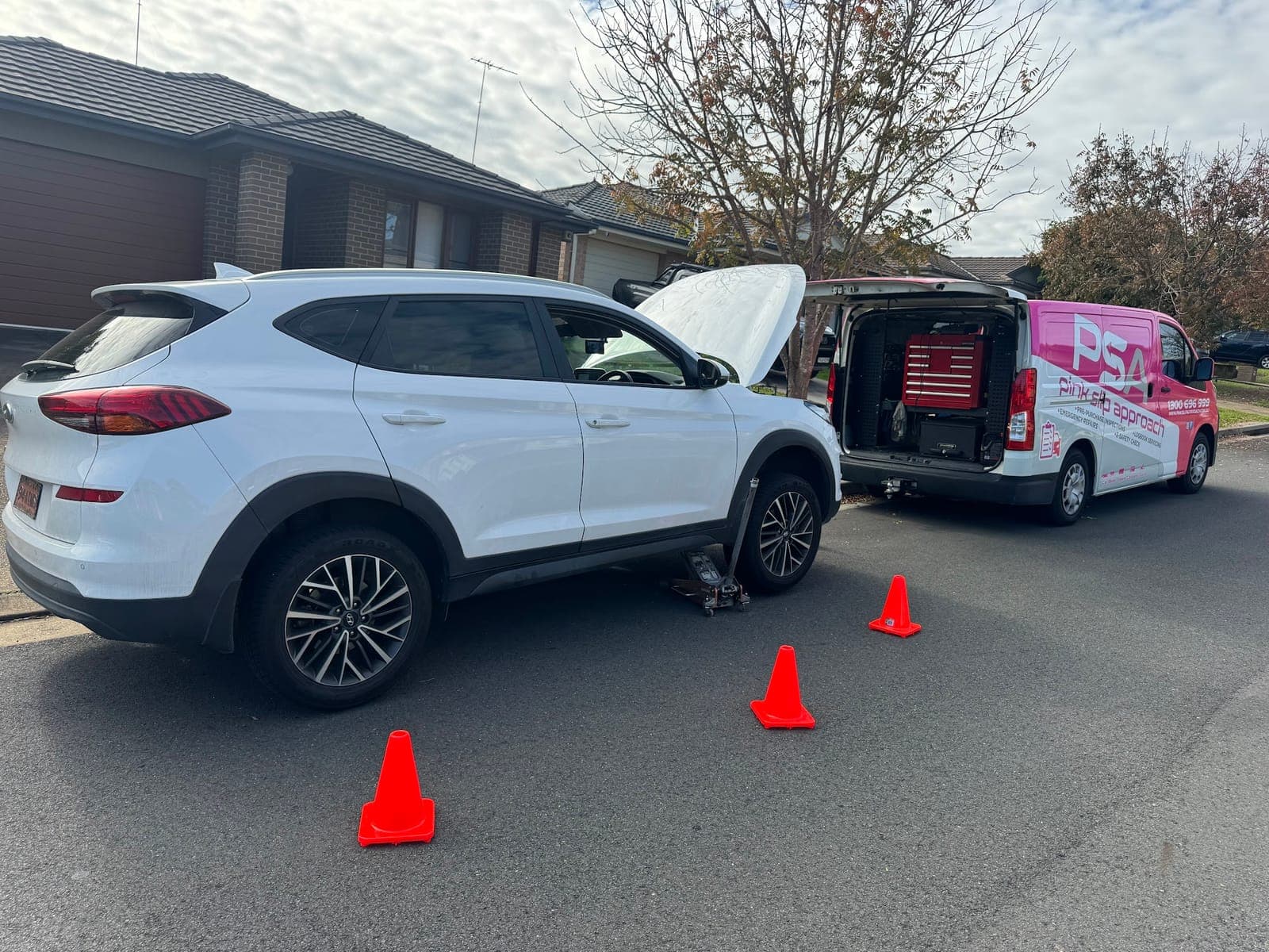A white Hyundai SUV on a jack with its bonnet up in a quiet street, a PSA van open behind it with its test gear inside